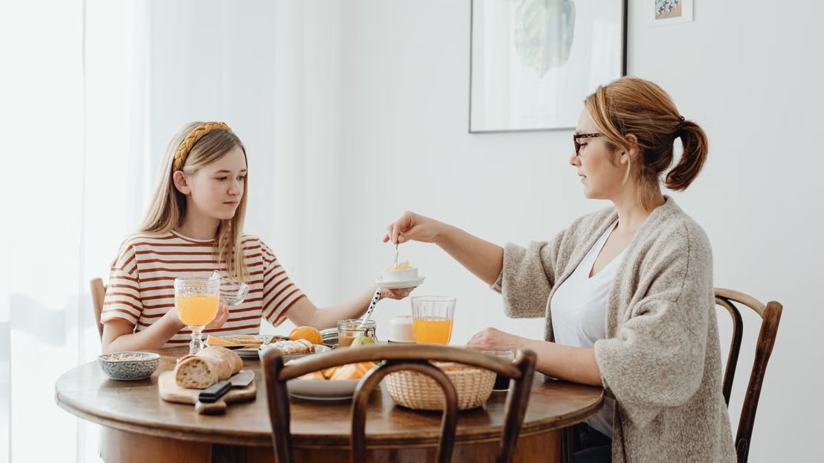 A mother and daughter share a peaceful breakfast with orange juice and bread at a wooden table.